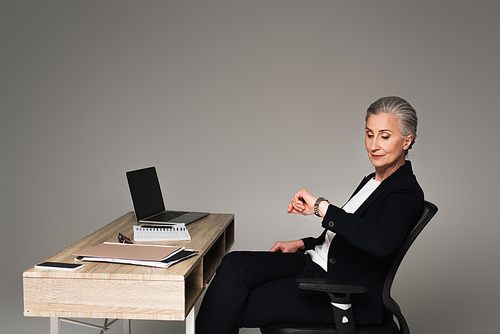 Businesswoman looking at wristwatch near devices on table on grey background
