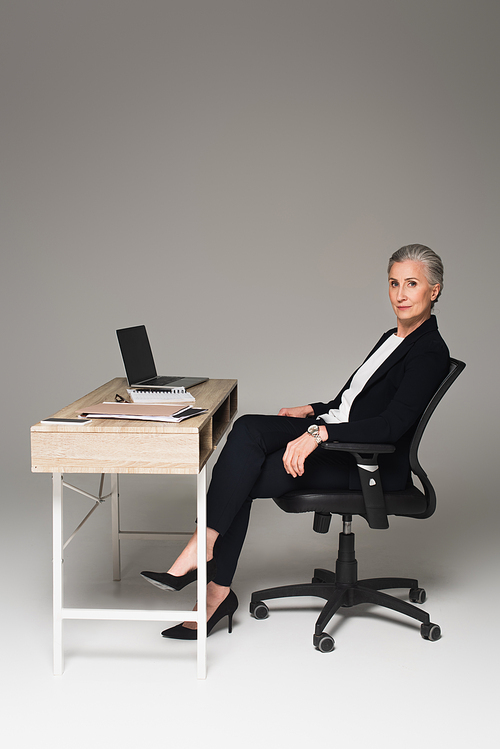 Mature businesswoman sitting near gadgets and papers on table on grey background