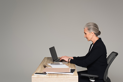 Side view of businesswoman using laptop near papers isolated on grey