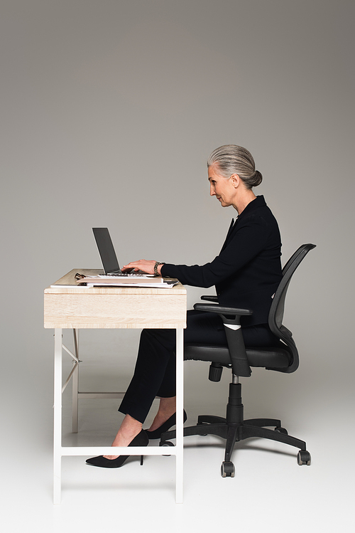 Side view of mature businesswoman using laptop near documents on table on grey background