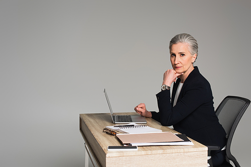 Mature businesswoman  near gadgets and papers on table isolated on grey