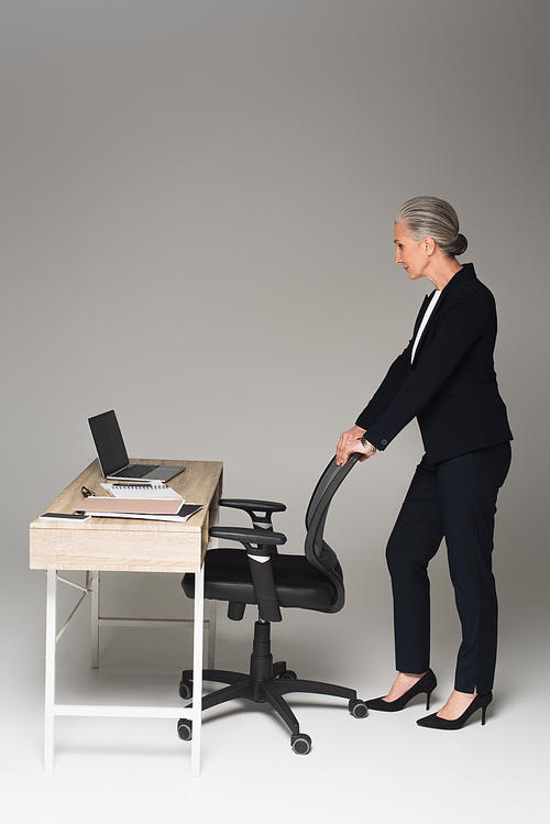 Side view of mature businesswoman standing near office chair and working table on grey background