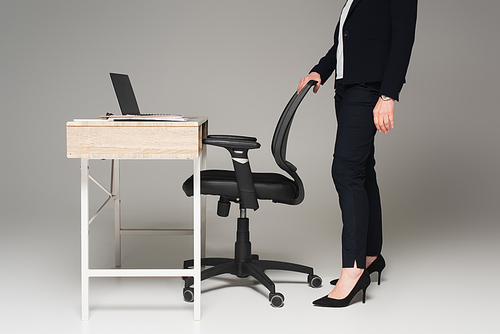 Cropped view of businesswoman standing near office chair and laptop on table on grey background