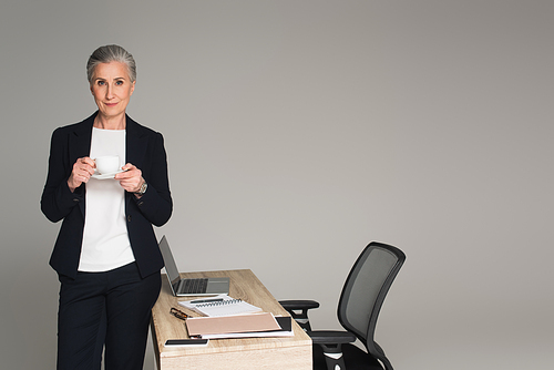 Mature businesswoman holding cup near papers and gadgets on table isolated on grey