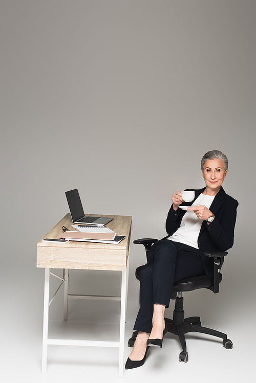 Mature businesswoman holding cup near gadgets and paperwork on table on grey background
