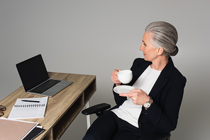 Middle aged woman with cup of coffee sitting near laptop and notebook on table isolated on grey