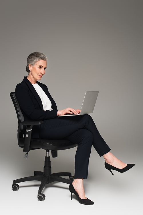 Mature businesswoman using laptop on office chair on grey background