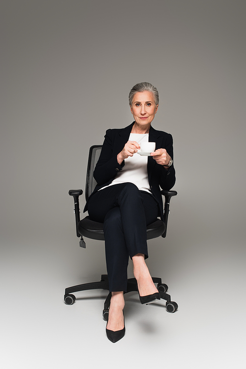 Businesswoman in formal wear holding cup of coffee on office chair on grey background