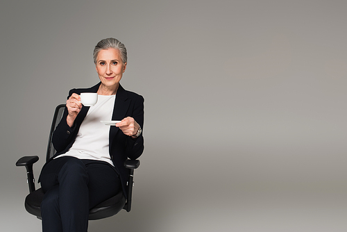 Mature businesswoman holding cup of coffee on office chair isolated on grey