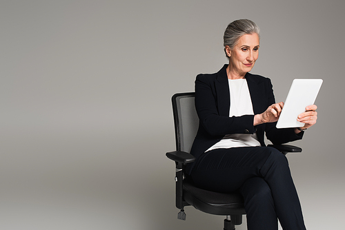 Grey haired businesswoman using digital tablet on office chair isolated on grey