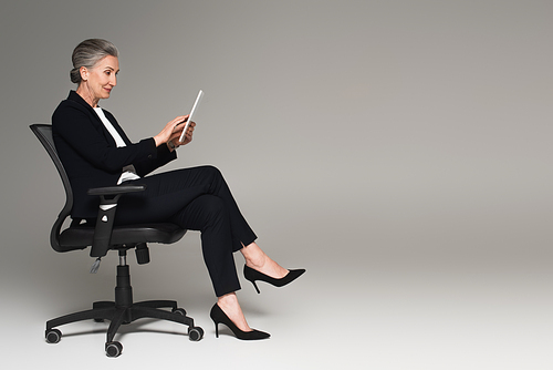 Mature businesswoman using digital tablet on office chair on grey background with copy space