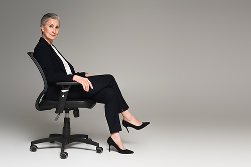 Middle aged businesswoman with crossed legs  on office chair on grey background
