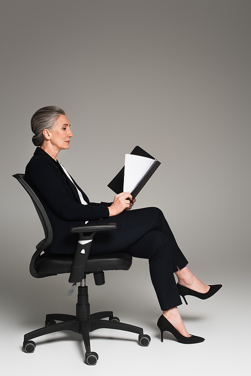 Side view of mature businesswoman looking at documents on office chair on grey background