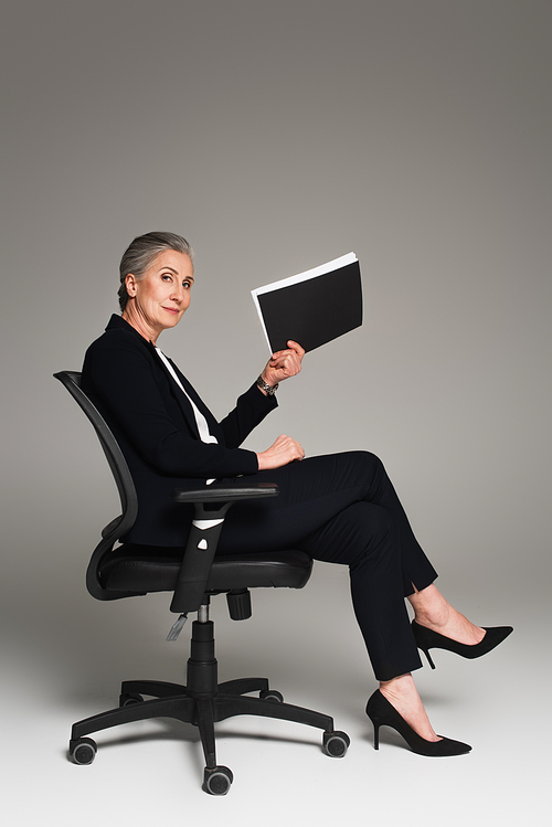 Mature businesswoman holding paper folder on office chair on grey background