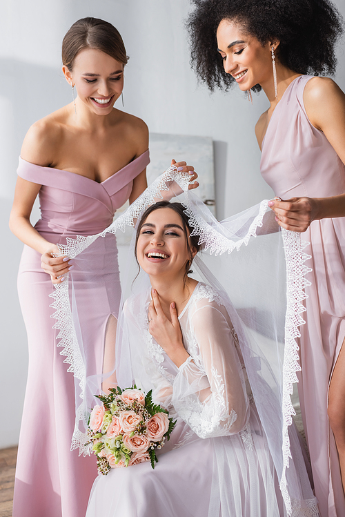 happy multicultural women covering laughing bride with veil in bedroom
