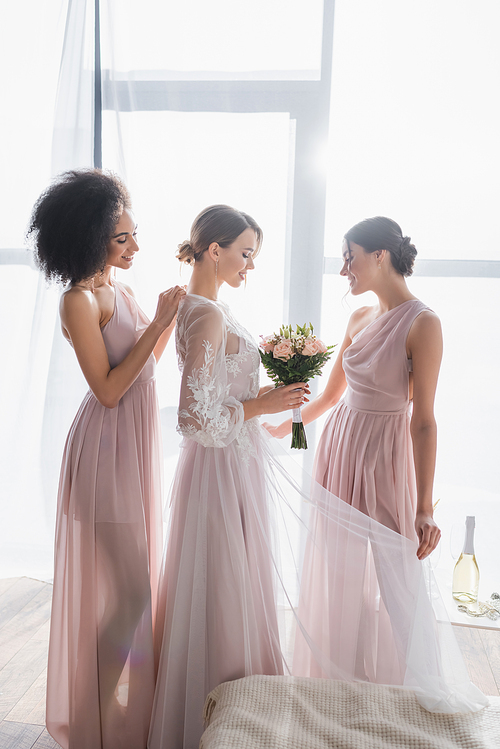 pretty bride holding wedding bouquet while interracial bridesmaids adjusting her dress on sunny morning