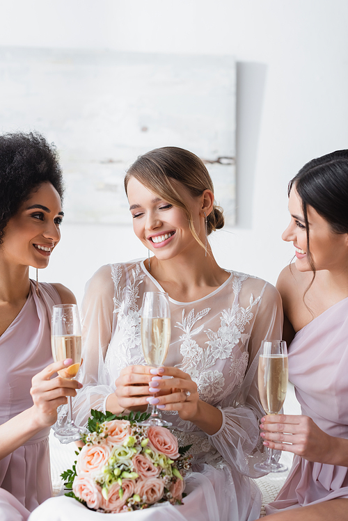 multicultural bridesmaids looking at happy bride while holding champagne glasses