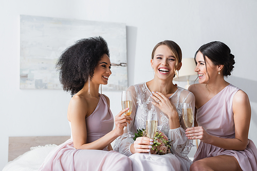 young bride laughing near interracial friends holding champagne glasses in bedroom
