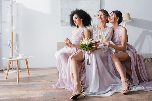 elegant interracial bridesmaids holding champagne glasses while sitting near bride in bedroom