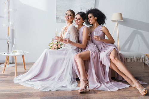 elegant bride with interracial bridesmaids sitting with champagne glasses in bedroom