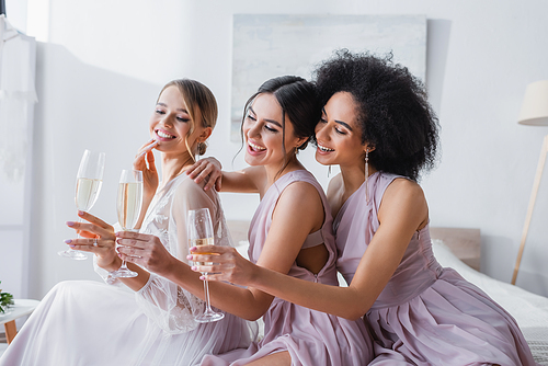 joyful bride and bridesmaids sitting in bedroom with champagne glasses