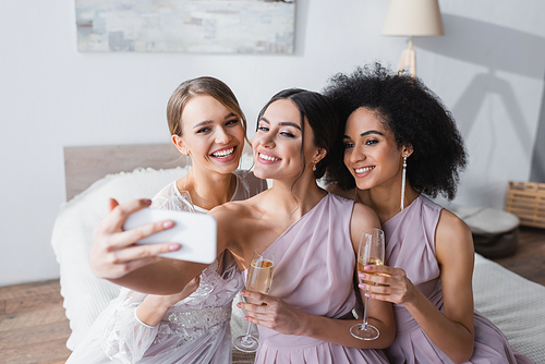cheerful woman taking selfie with bride and african american friend in bedroom