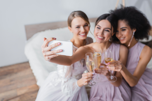 joyful woman taking selfie with bride and african american friend on blurred background