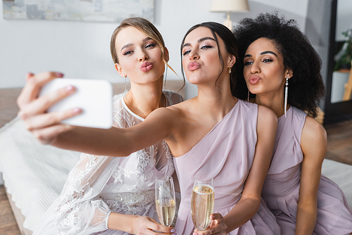 pretty bride with multicultural bridesmaids blowing air kisses while taking selfie on blurred foreground