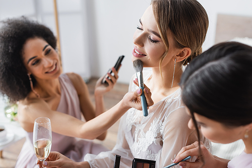 young bride holding champagne glass while multicultural bridesmaids applying makeup, blurred background