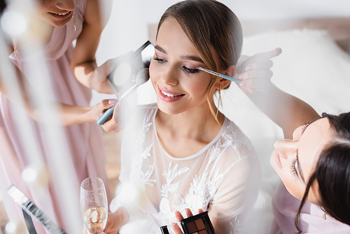 interracial women doing makeup to young bride holding champagne glass on blurred foreground