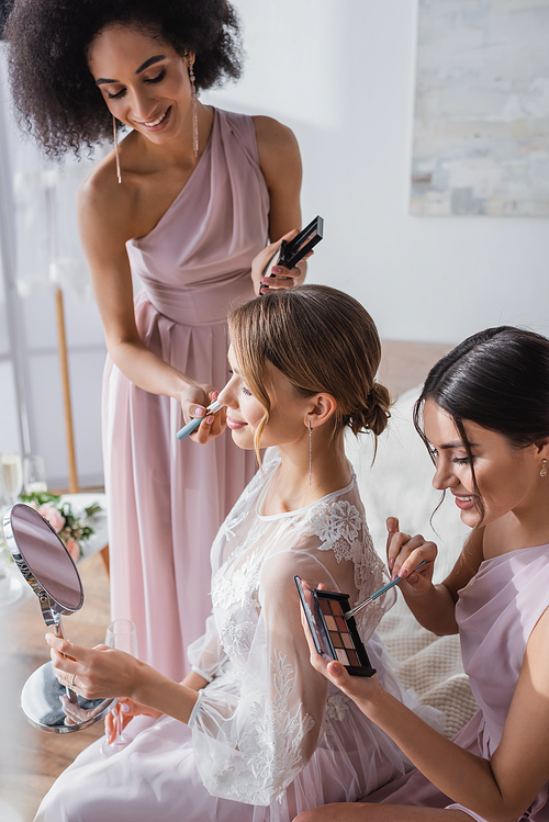 young bride looking in mirror while interracial women applying makeup