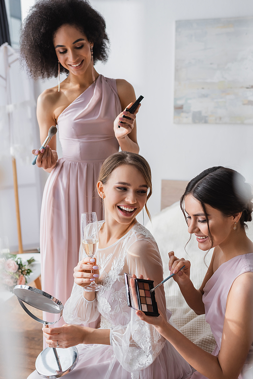 laughing bride holding champagne and mirror near interracial friends with decorative cosmetics