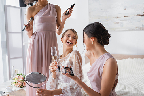 excited bride laughing near interracial friends holding decorative cosmetics