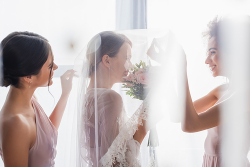 side view of happy interracial bridesmaids near smiling bride in veil on blurred foreground