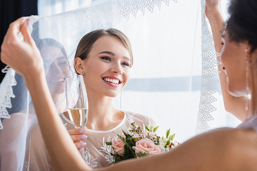 african american bridesmaid holding veil of happy bride with champagne glass and bouquet