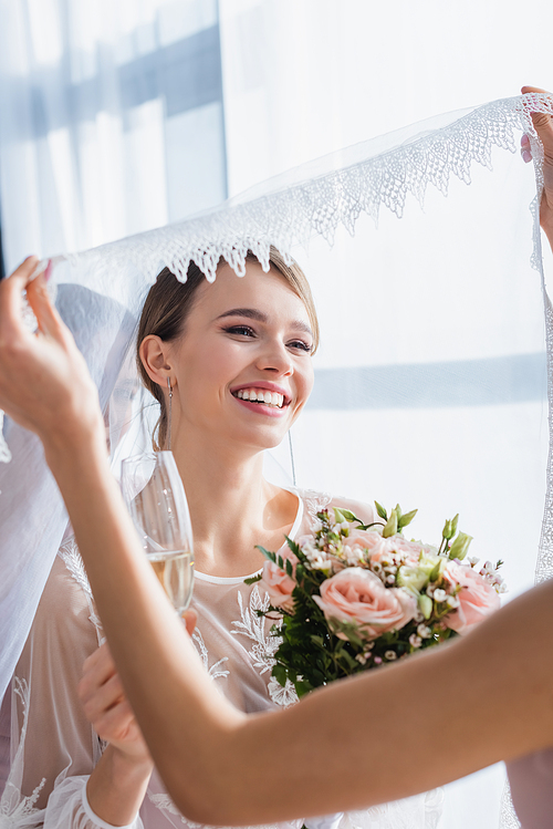 bridesmaid holding veil of joyful bride with wedding bouquet and champagne