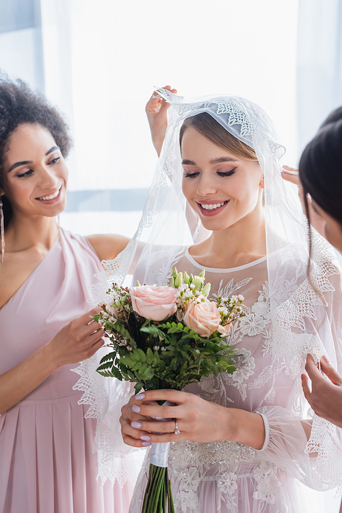 smiling bride holding wedding bouquet near interracial bridesmaids