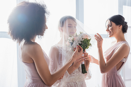 bride holding wedding bouquet with closed eyes while multicultural bridesmaids covering her with veil
