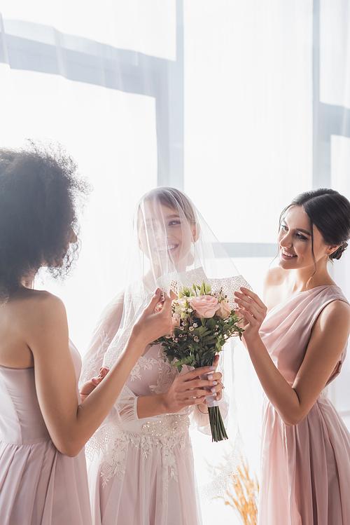 multicultural bridesmaids touching veil of smiling bride holding wedding bouquet
