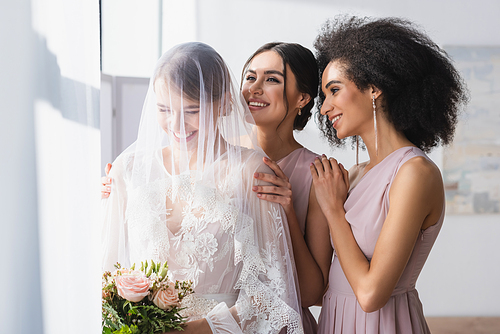 happy bride holding wedding bouquet near smiling interracial bridesmaids