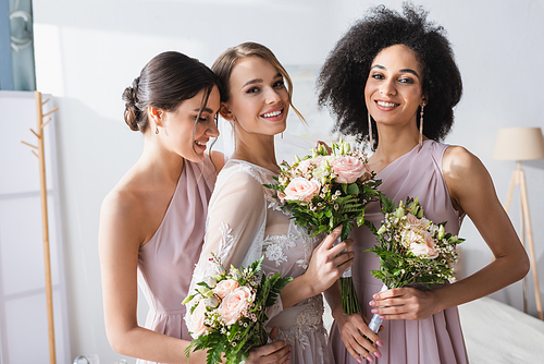 pretty bride together with interracial bridesmaids holding wedding bouquets in bedroom