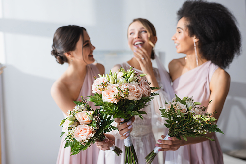 laughing bride and multicultural bridesmaids holding wedding bouquets on blurred background