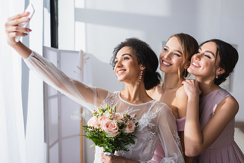 young african american bride taking selfie with cheerful bridesmaids at home