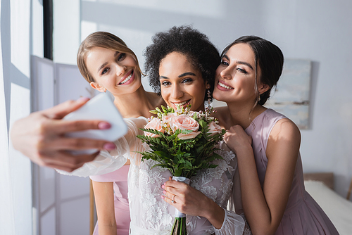 african american bride taking selfie with wedding bouquet and bridesmaids, blurred foreground