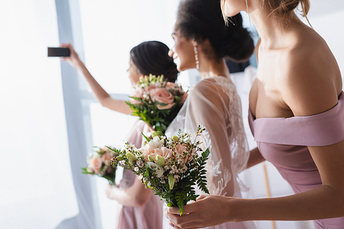 bride taking selfie with interracial bridesmaids holding wedding bouquets, blurred background