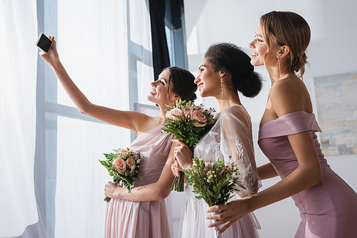 african american bride with bridesmaids holding wedding bouquets while taking selfie on smartphone