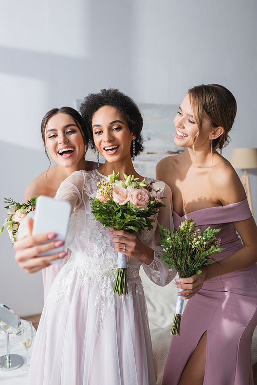 african american bride taking selfie with cheerful bridesmaids at home
