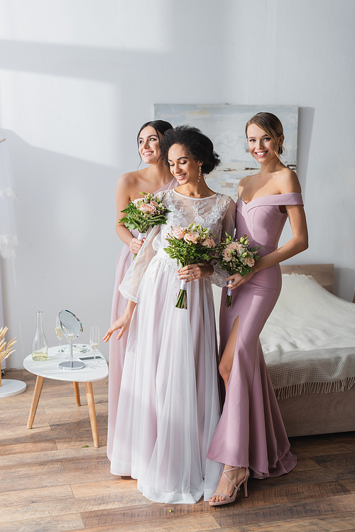 elegant african american bride with happy bridesmaids standing with wedding bouquets in bedroom