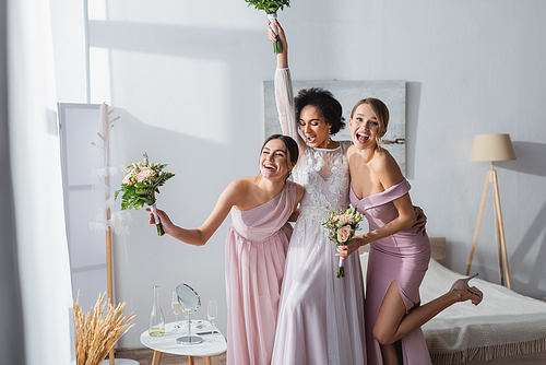 excited african american bride with bridesmaids holding wedding bouquets while having fun in bedroom