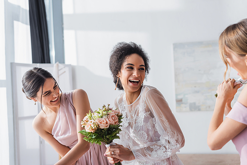 excited african american bride holding wedding bouquet and laughing near bridesmaids
